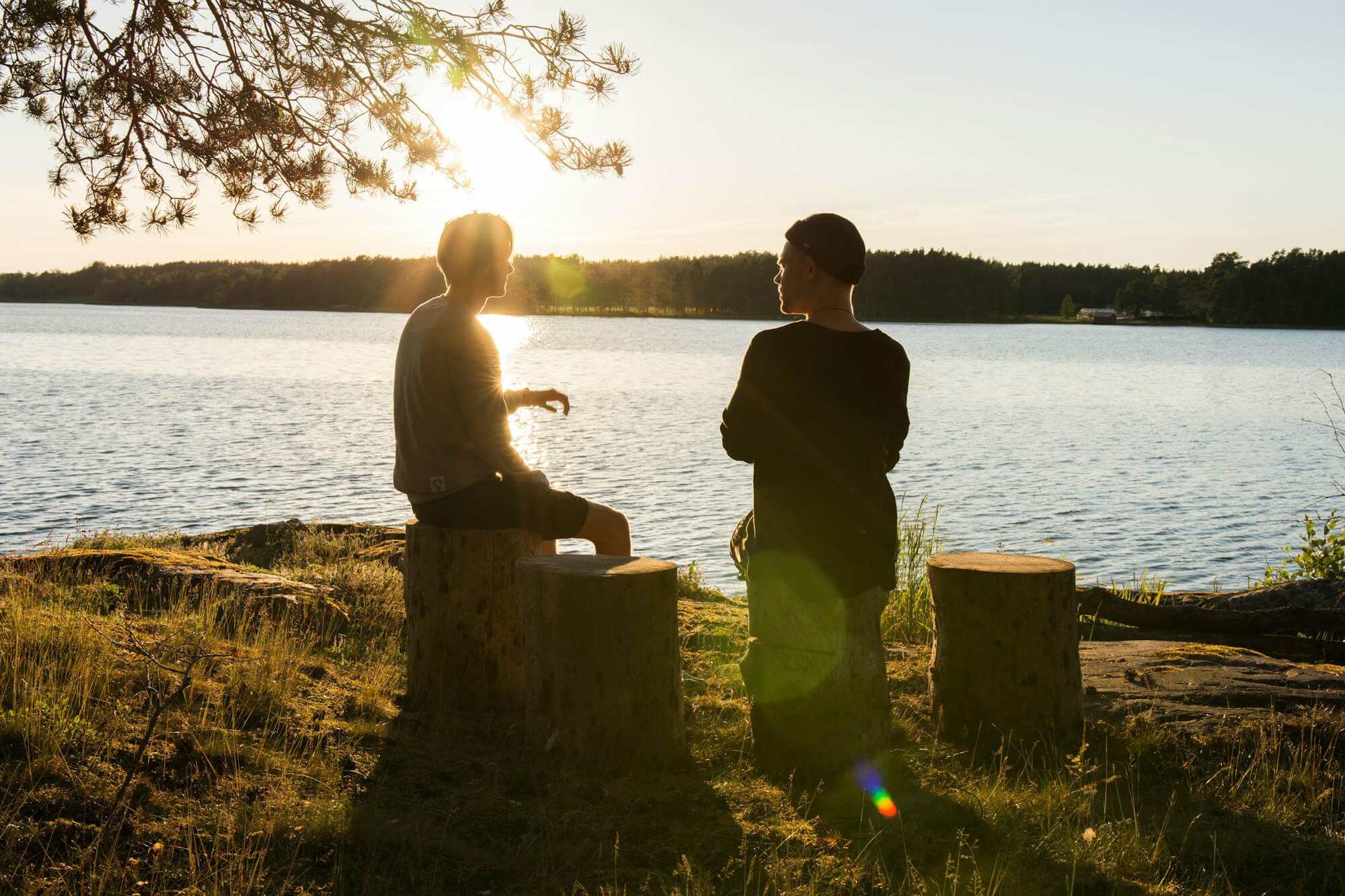 Zwei Männer sitzen im Gegenlicht auf Holzstümpfen am Ufer vor einem See